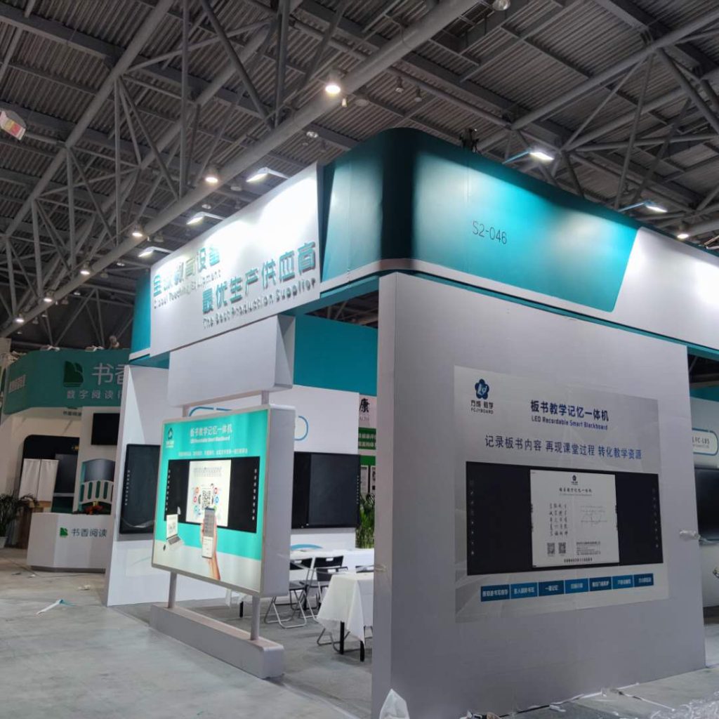 Real photo of an exhibition booth with teal accents and white walls, displaying large screens with educational content and a QR code.