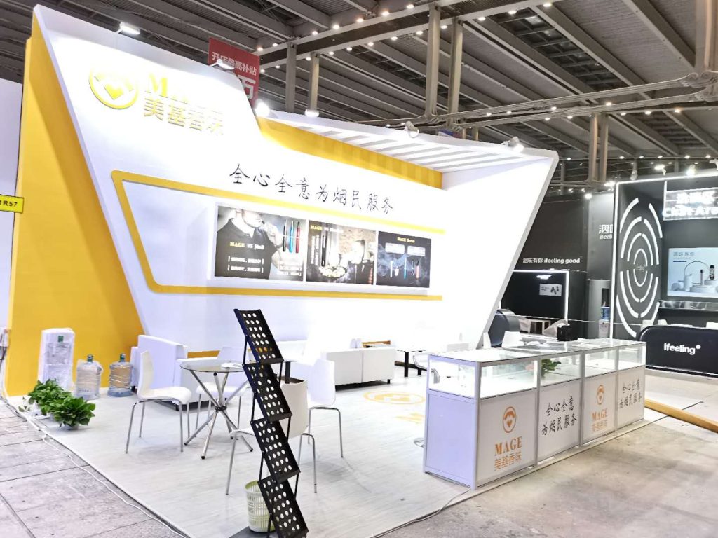 A white and yellow trade show booth in a large hall, featuring display cases, a literature rack, and a small seating area.