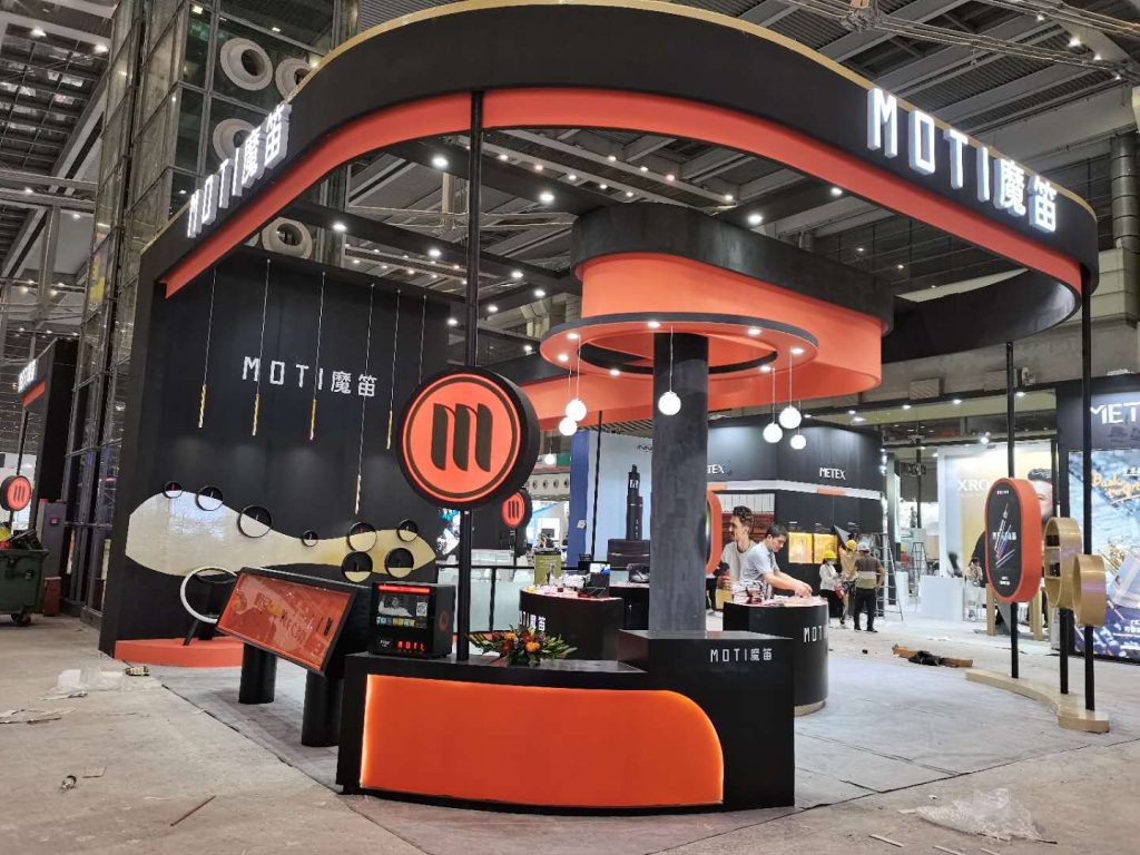Exhibition booth with black and orange design, featuring display counters and circular signage, with workers nearby at a trade show.