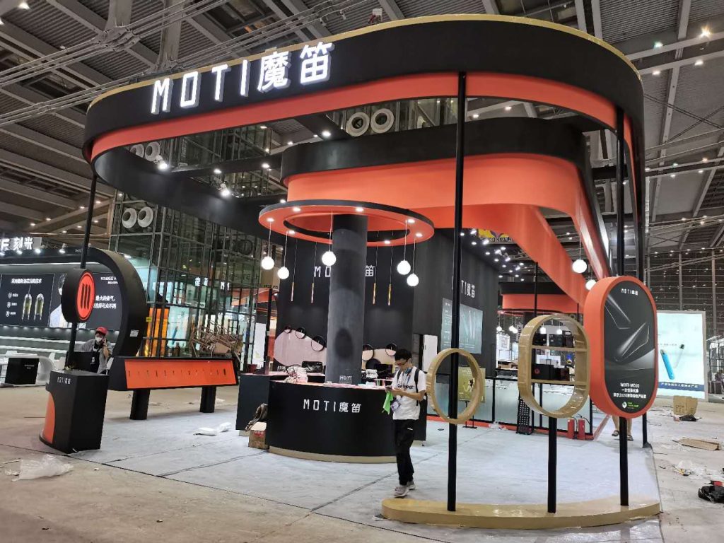 Exhibition booth in black and orange, under construction, with display units and a person wiping a counter. Industrial ceiling visible.
