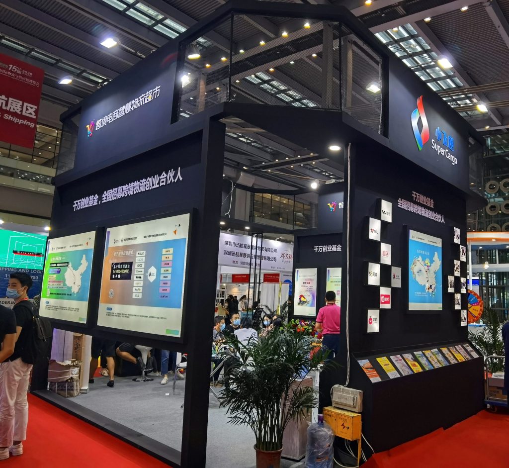 A trade show booth with various display screens showing maps and charts, pamphlets on a wall, and several people walking around.