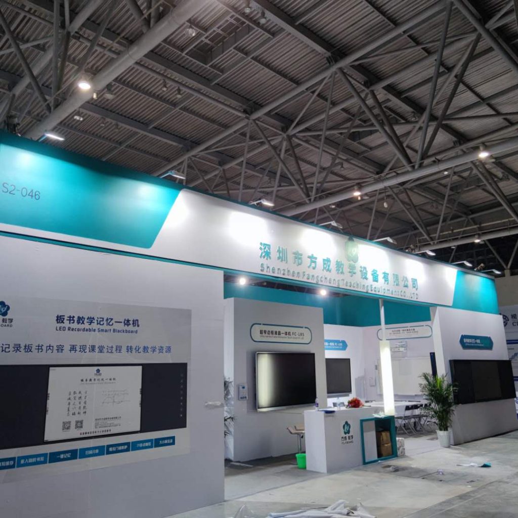 Real photo of a spacious exhibition booth with several display screens showing content, a small plant, and a prominent overhead banner.