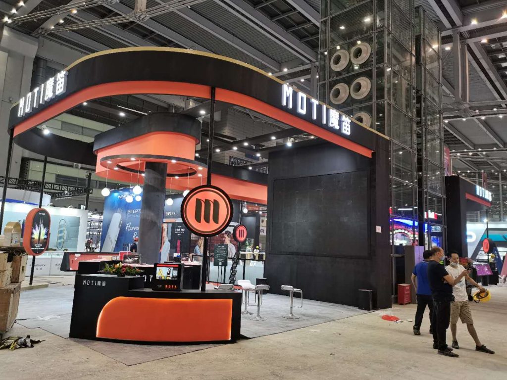 Exhibition booth with black and orange counters and stools. People stand nearby, with other exhibit halls visible in the background of a trade show.
