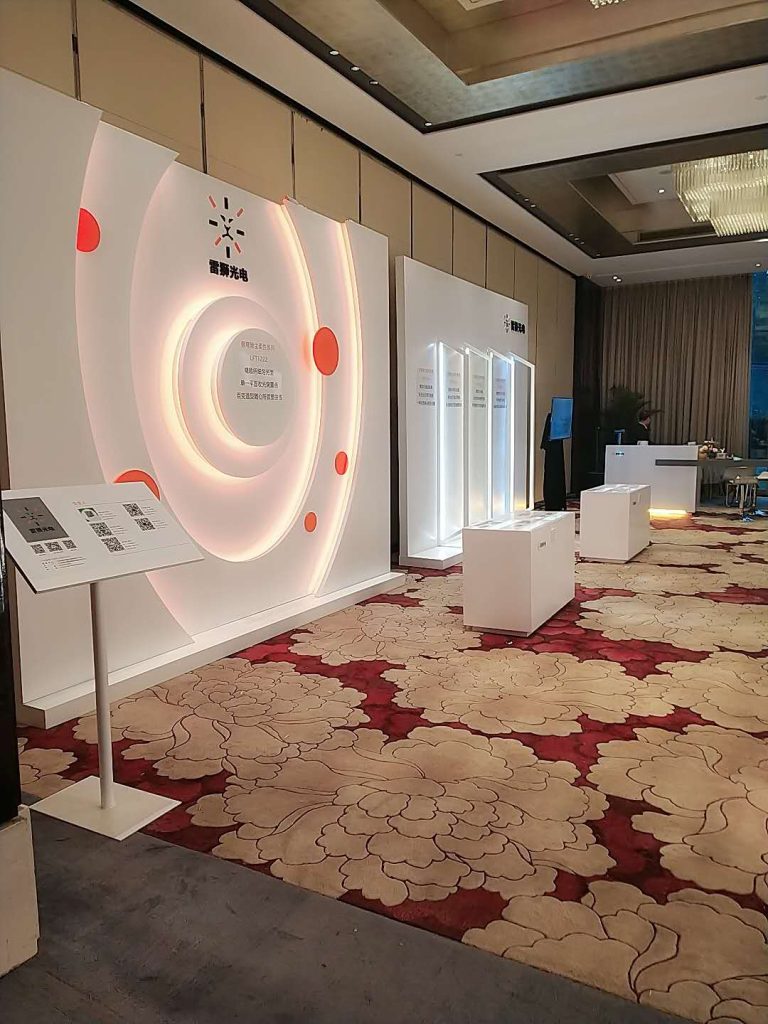 A conference hall with a white exhibit wall featuring illuminated concentric circles and red accents, a display stand, and patterned carpet.
