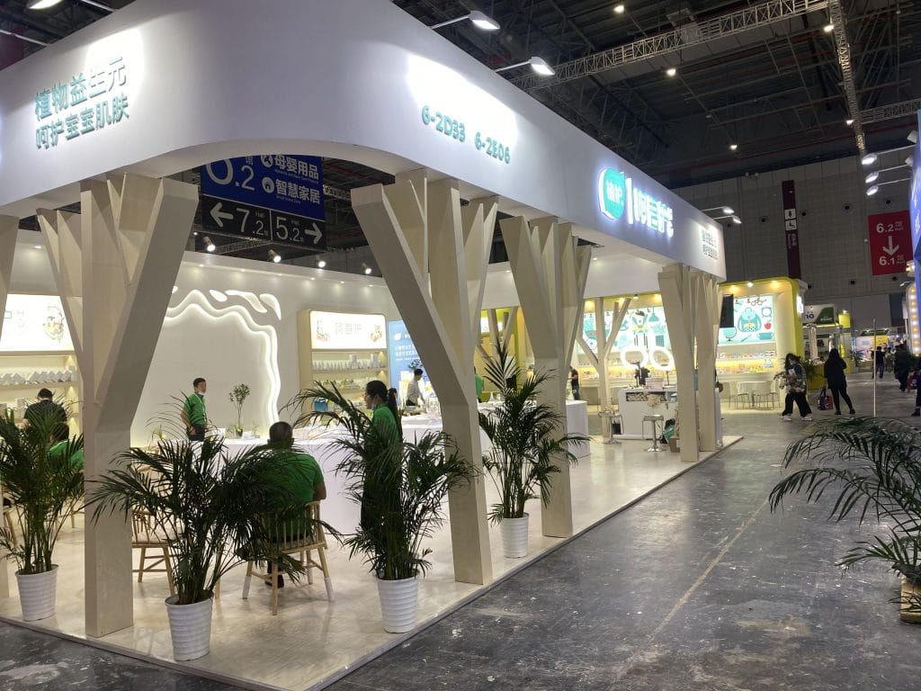 Busy trade show booth with white canopy, wooden pillars, plants, and visitors.