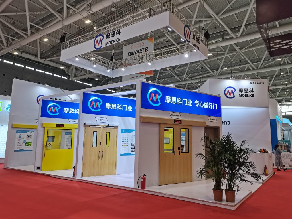 A wide view of a modern exhibition booth featuring several industrial and wooden doors, a large potted plant, and overhead signage in a large hall.