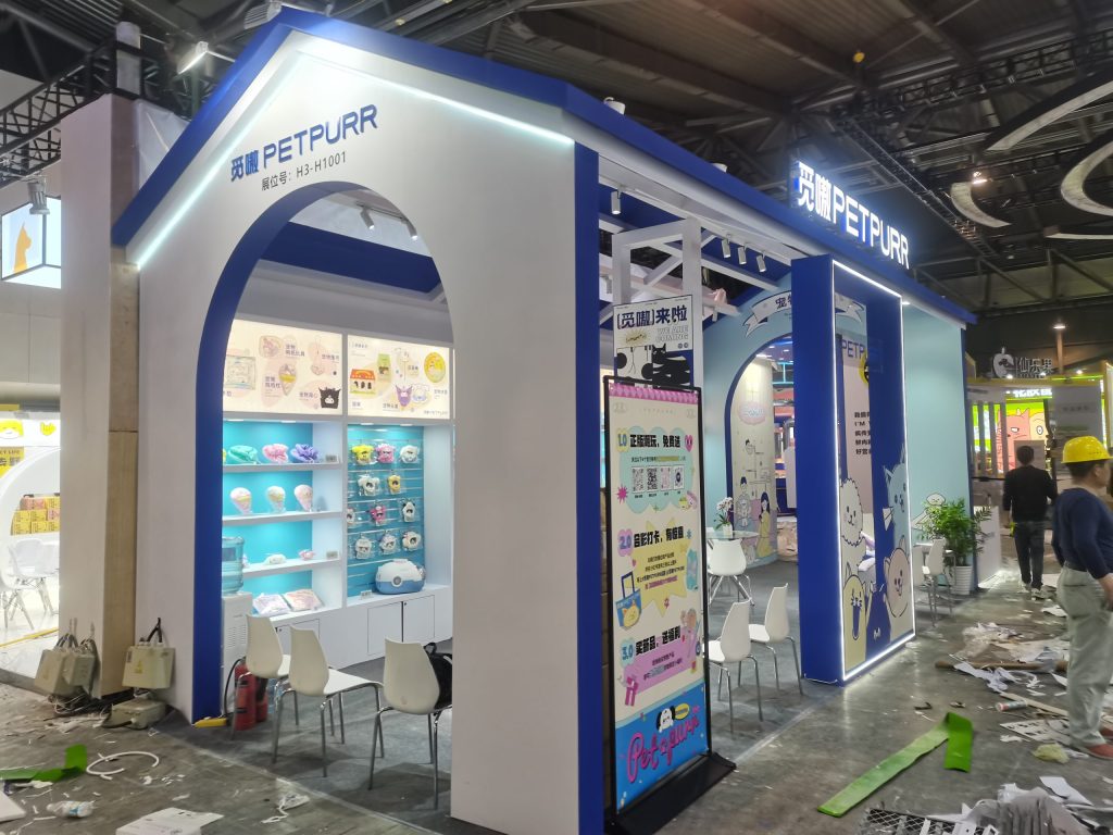House-shaped trade show booth under construction. Shelves are filled with products, debris on the floor, and a worker in a hard hat.