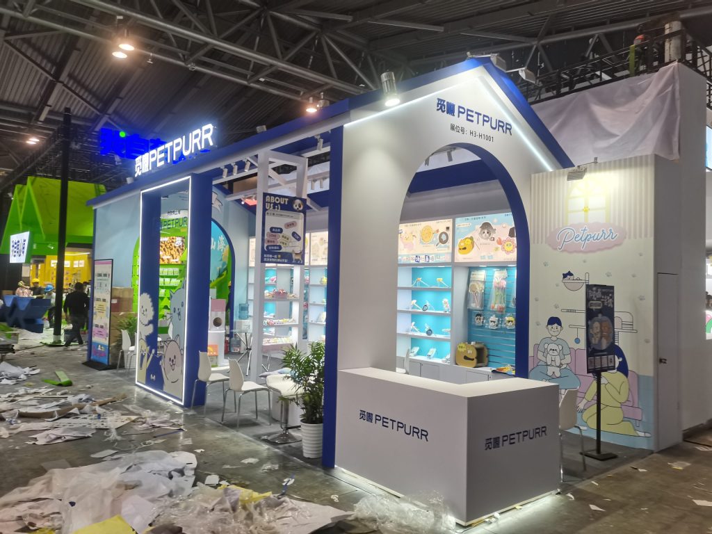 House-shaped trade show booth under construction. A white reception counter is visible, with debris and plastic on the floor.