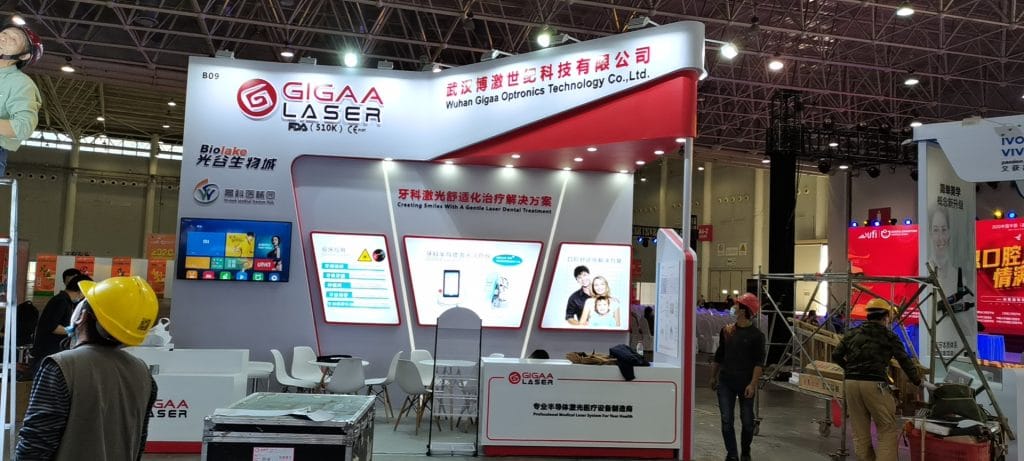 Two photos of an active red and white exhibition booth with digital screens, visitors, and staff members present.