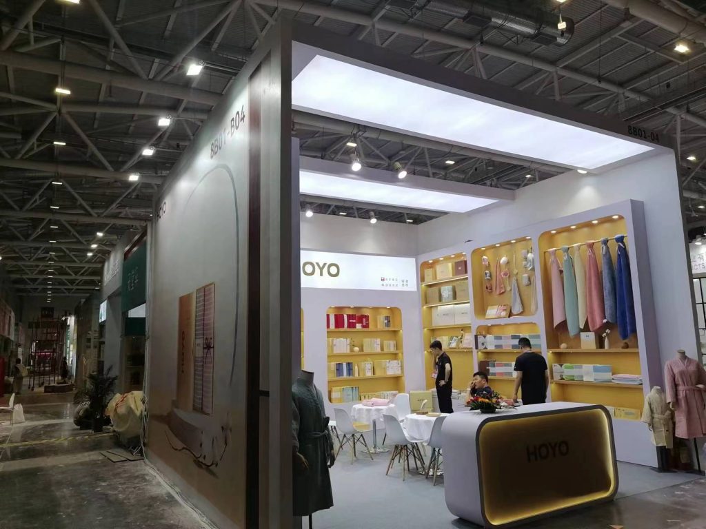 A wide shot of an exhibition booth in a large hall, showing display shelves, a reception counter, and a seating area.