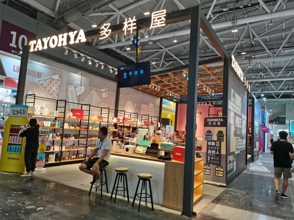 Exhibition booth with shelves displaying diverse products. A man in a mask sits at a counter with stools. Another person stands nearby.