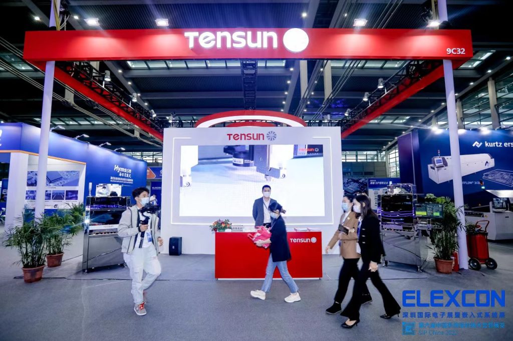 Busy exhibition hall with a prominent red and white booth. People wearing masks walk past a large screen displaying industrial machinery.