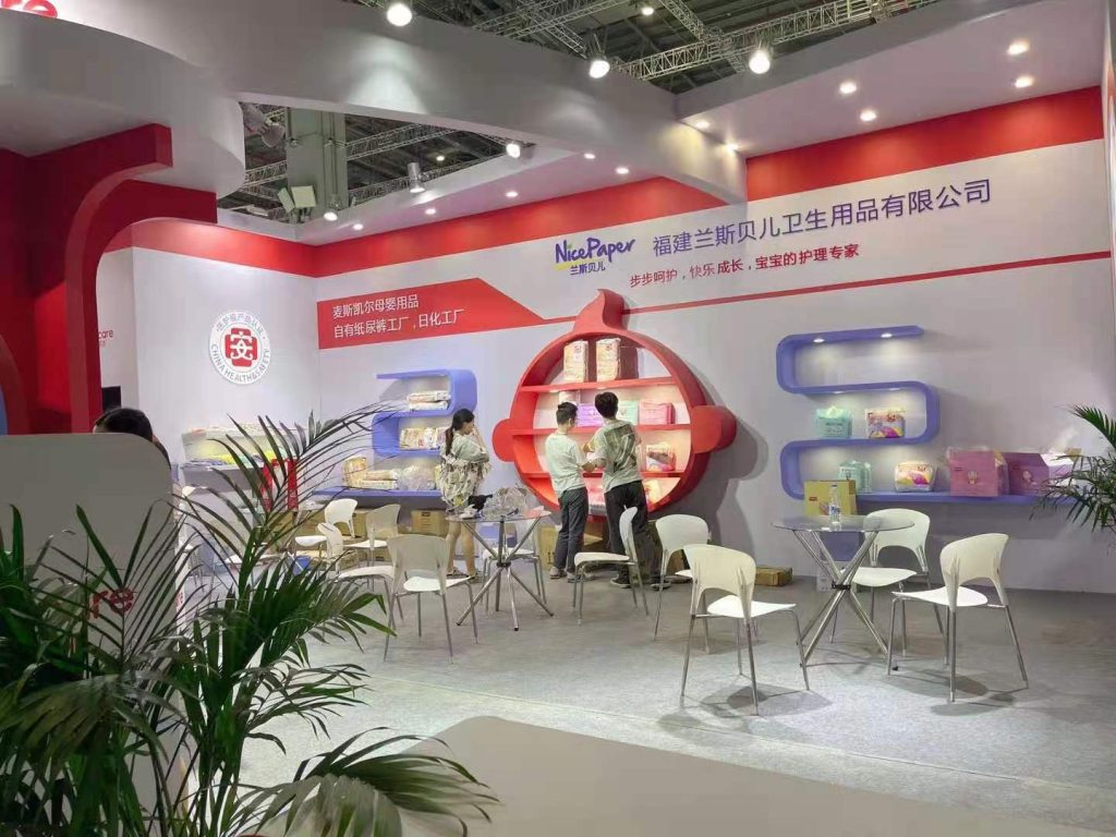 A brightly lit trade show booth with red and white walls, displaying packaged products on shelves. Several people mingle around tables and chairs.