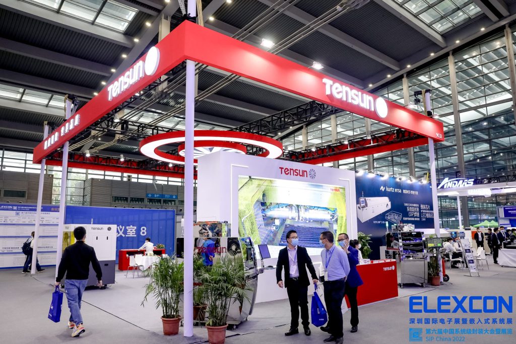 A spacious exhibition hall with a large red and white booth. Attendees, some masked, walk and gather. Green potted plants decorate the space.