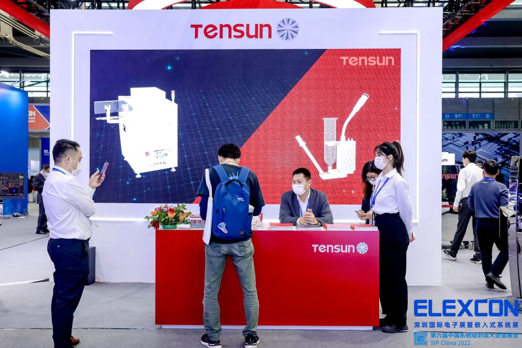Masked attendees interact at a red reception counter inside an exhibition booth. A large screen behind them displays white industrial equipment designs.