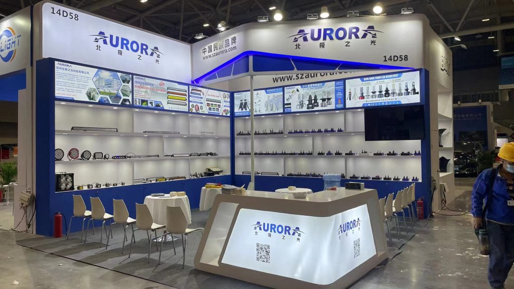 A blue and white trade show booth displaying rows of automotive lighting products on shelves, with a reception desk and tables for visitors.