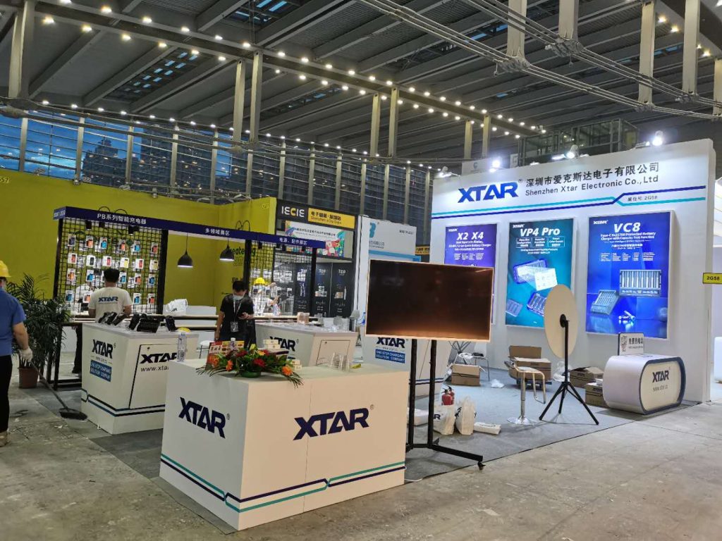 A large exhibition booth with white counters, display panels, and a dark screen on a stand. People walk around the hall.