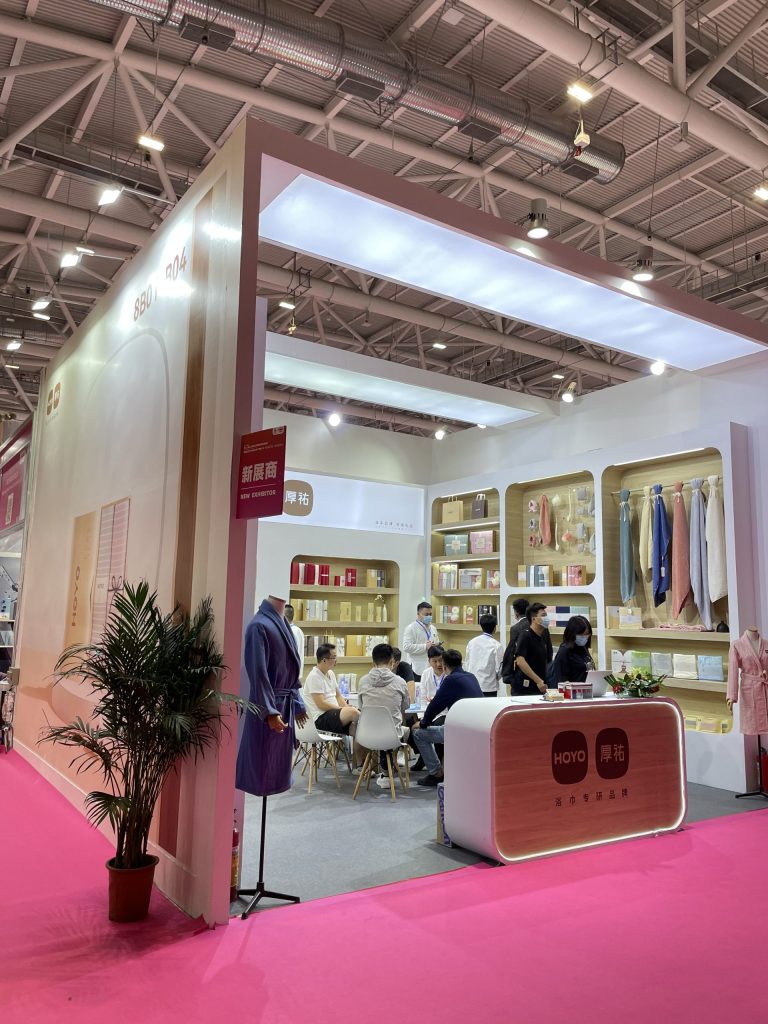 Busy exhibit booth with attendees seated at a table, products displayed on shelves, and bathrobes on mannequins. A potted plant is in the foreground.