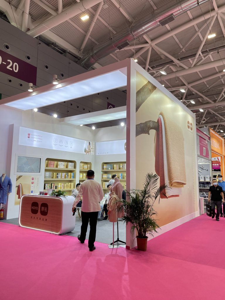 Man walking past an exhibit booth with a large graphic of towels, a display bathrobe, and a potted plant on a pink carpeted floor.