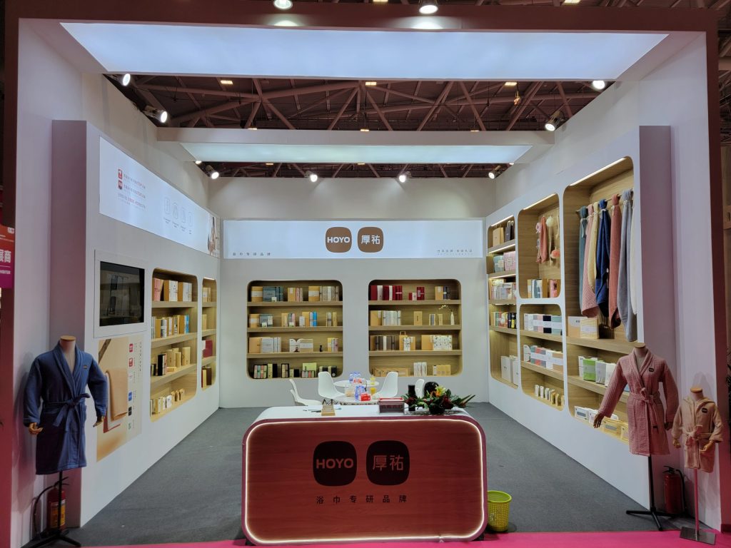Interior view of an exhibit booth featuring multiple shelves filled with packaged products, a central counter, and two bathrobes on display.