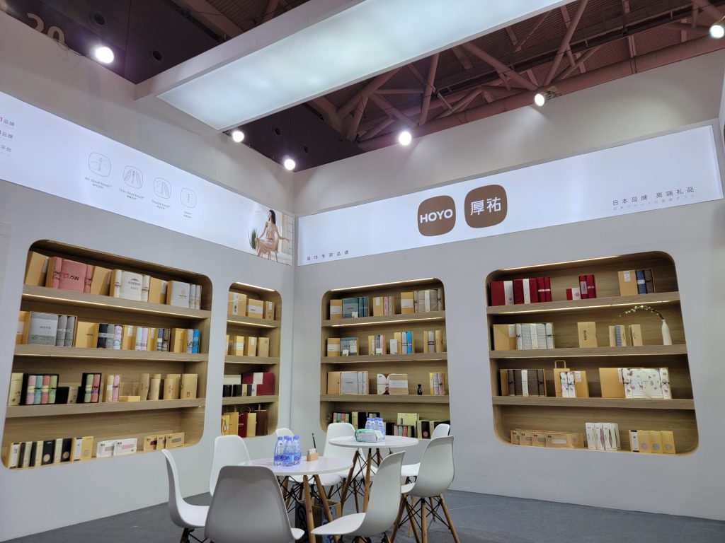 Exhibit booth interior with several people sitting at tables, surrounded by walls with display shelves full of various packaged items.