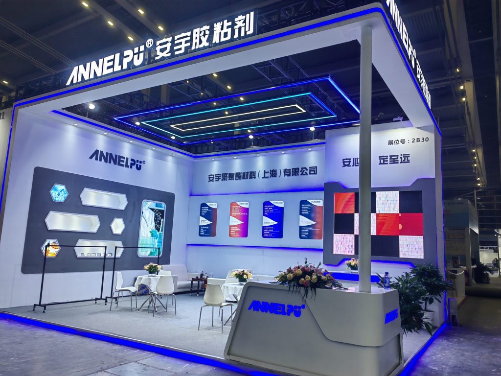 A brightly lit trade show booth with white and blue neon accents, display panels, two seating areas, and a reception desk with flowers.