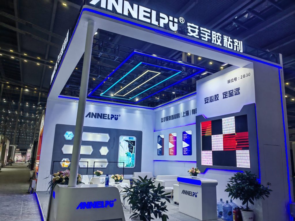 A brightly lit trade show booth with white and blue neon accents, display panels, seating, a reception desk with flowers, and potted plants.