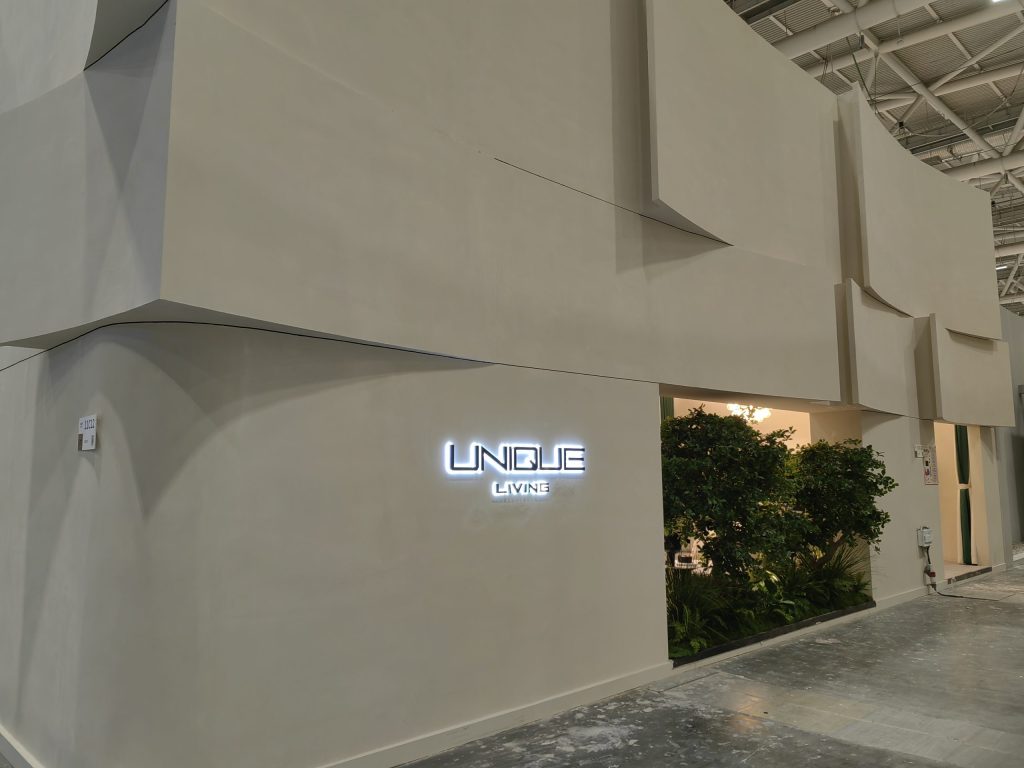 Exhibition booth with glowing "UNIQUE LIVING" sign, an open entrance revealing lush green plants, and light-colored modular walls.