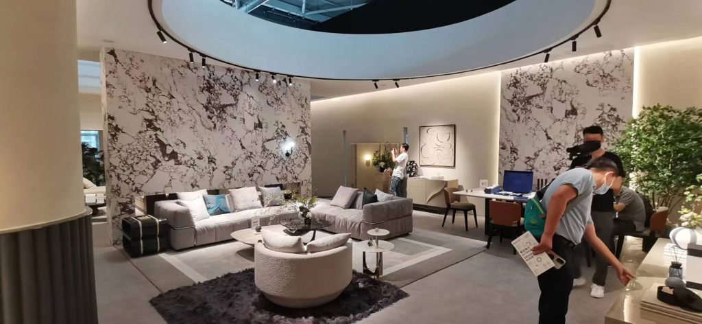 Luxurious living room with a grey sectional sofa, white marble accent wall, a cream circular chair, and several people browsing the furniture.