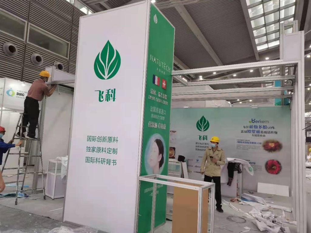 Workers assemble a white and green exhibition booth with leaf logos and Chinese text, featuring a person on a ladder and another standing nearby.