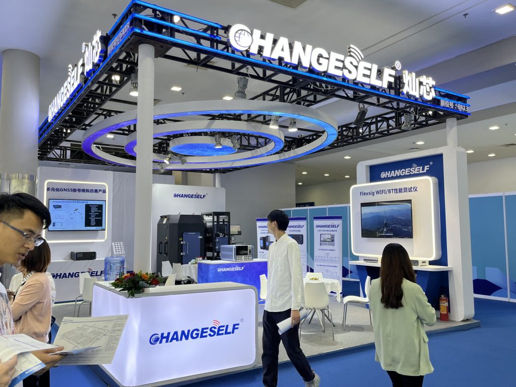 People browse a brightly lit exhibition booth with blue accents, multiple display screens, and a central counter.