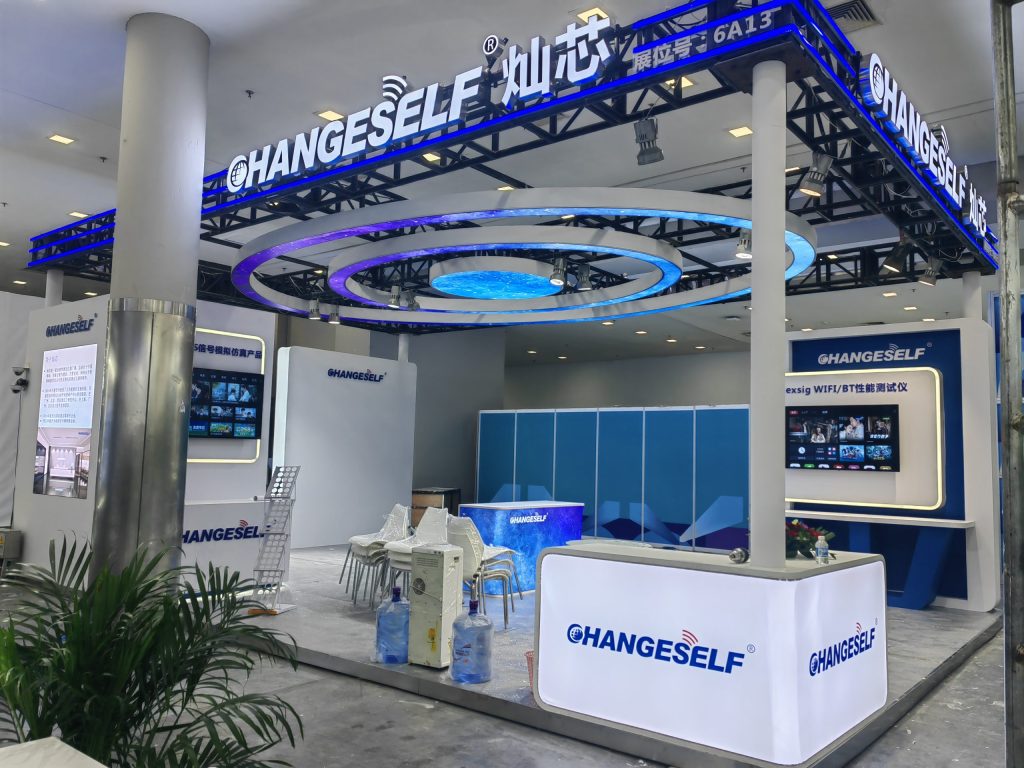 A clean, empty exhibition booth with blue and white illuminated structures, plants, and a reception counter.