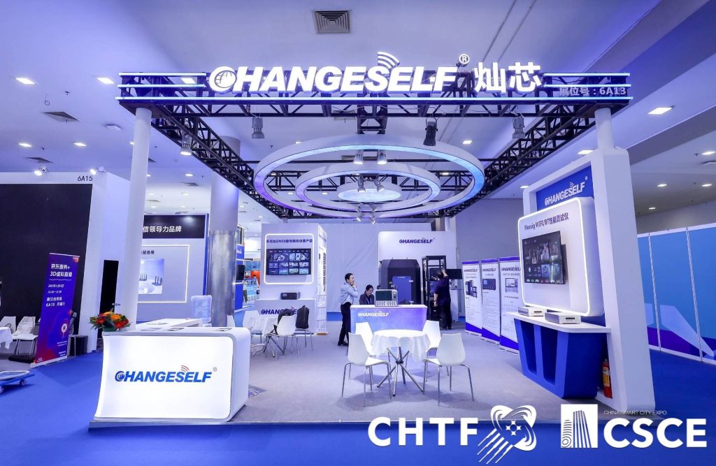 Visitors interact at a brightly lit exhibition booth with blue accents, multiple screens, and a small seating area.