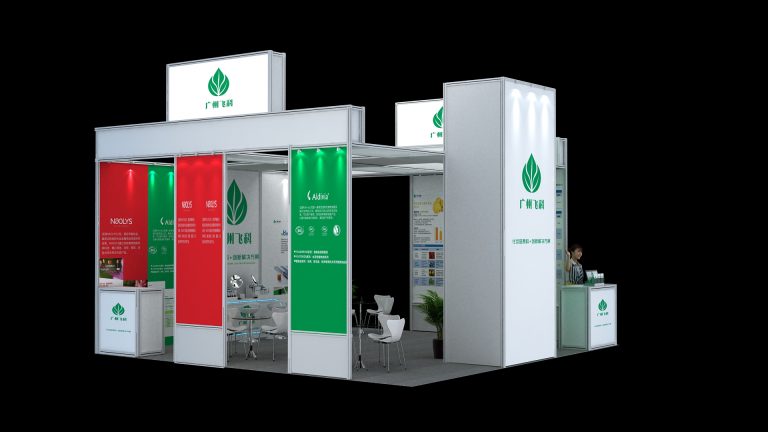 An exterior view of a large, open exhibition booth with white frames, red and green display panels, leaf logos, and a reception counter with a person.