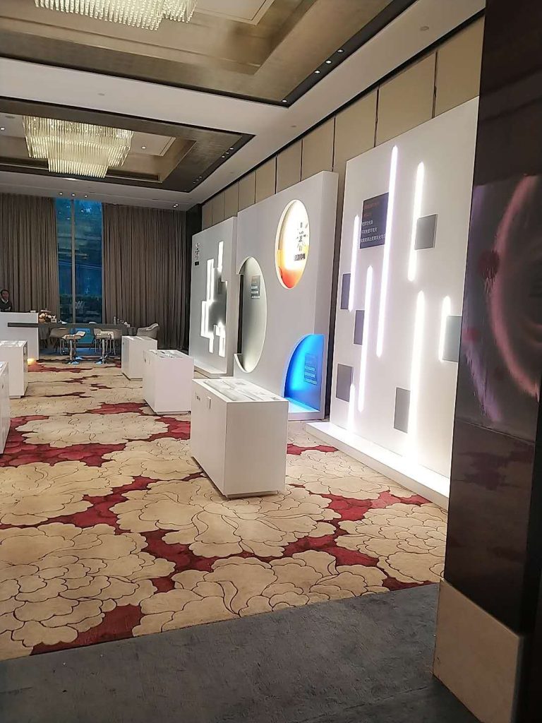 A conference hall with illuminated white exhibit walls, some with vertical lights and a blue panel, on a patterned red and beige carpet.