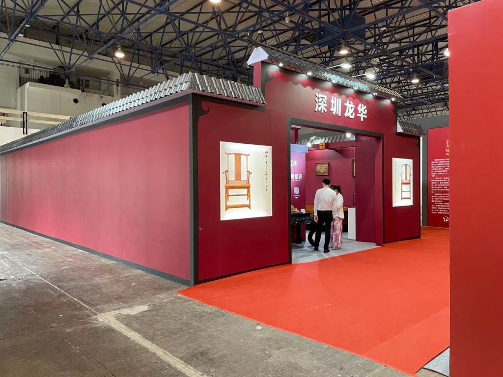 Exhibition booth with a red facade, traditional roof, and an arched entrance. Display cases show a wooden chair. Two people stand inside on a red carpet.