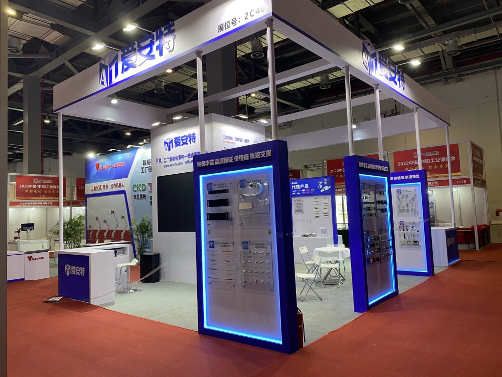 A white and blue trade show booth on a red carpet, featuring illuminated product displays, a reception desk, and a small seating area.