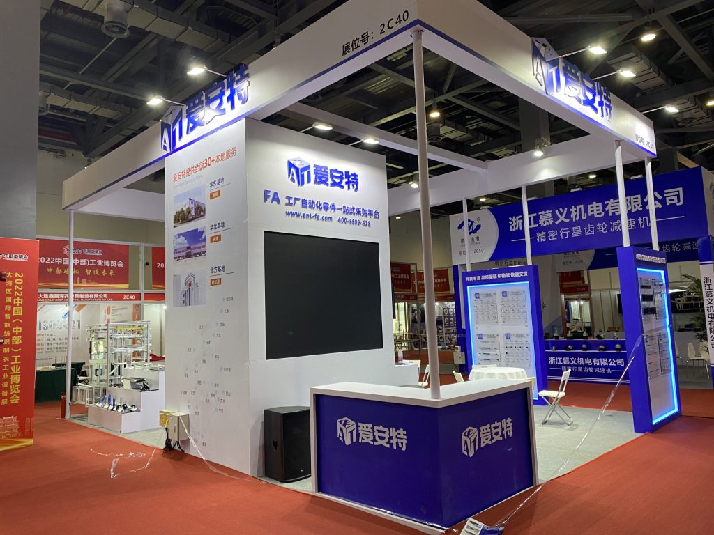 A white and blue trade show booth on red carpet, featuring a large screen, reception desk, and multiple product display boards.