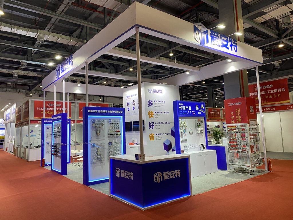 A wide view of a white and blue trade show booth with various product displays, a reception desk, and seating on a red carpet.