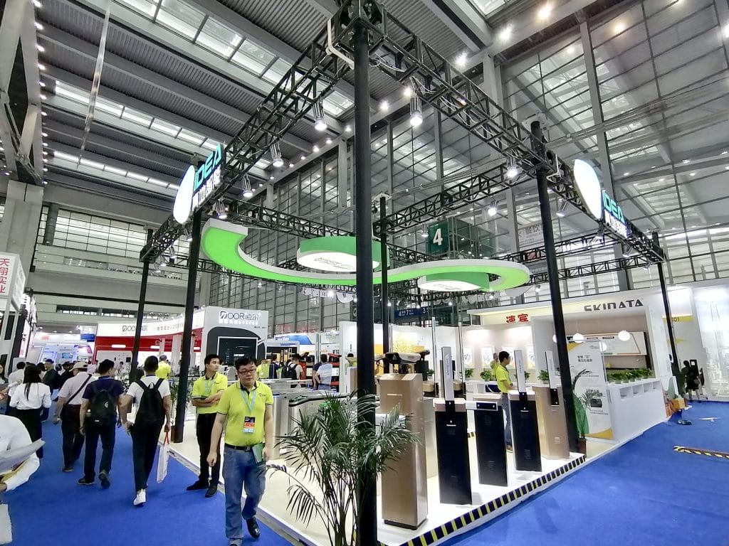 Trade show booth with black truss, green curved lights, and several dark-colored access control devices. People walk on a blue carpet in a large exhibition hall.