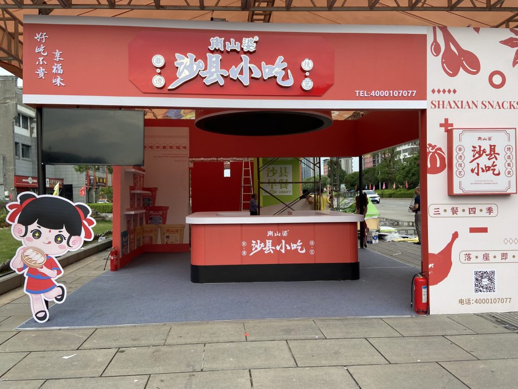 Outdoor red food stall under a tent, featuring a counter, screen, and large cartoon character. Construction workers are present.