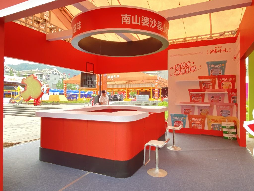 Inside a red food stall with a white counter, two stools, and shelves displaying packaged items. A person is visible behind the counter.