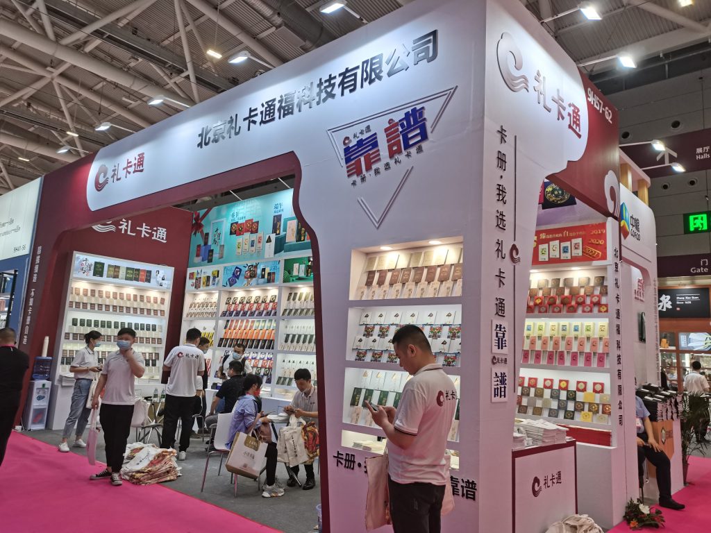 An exhibition booth with numerous small packages displayed on white shelves. Several people, some masked, browse products or use phones on a pink carpeted floor.