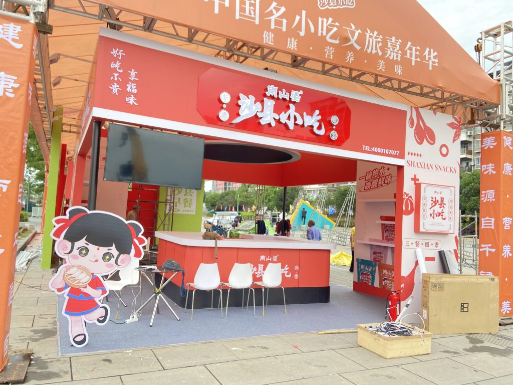 Outdoor red food stall with a serving counter, seating, and a large cartoon character cutout on a gray outdoor surface.
