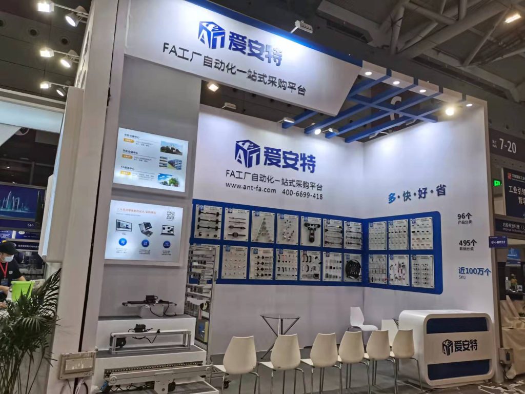 Exhibition booth with large blue and white signage. Displays of industrial parts line the back wall, with a long machine on a lower display. White chairs are arranged.