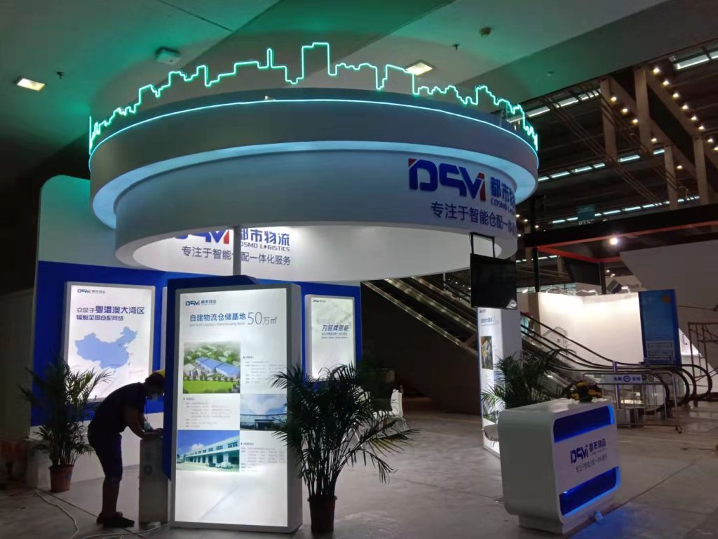 A real trade show booth, white and blue, with a green neon skyline. A person adjusts a lighted panel displaying a map. Escalators are in the background.