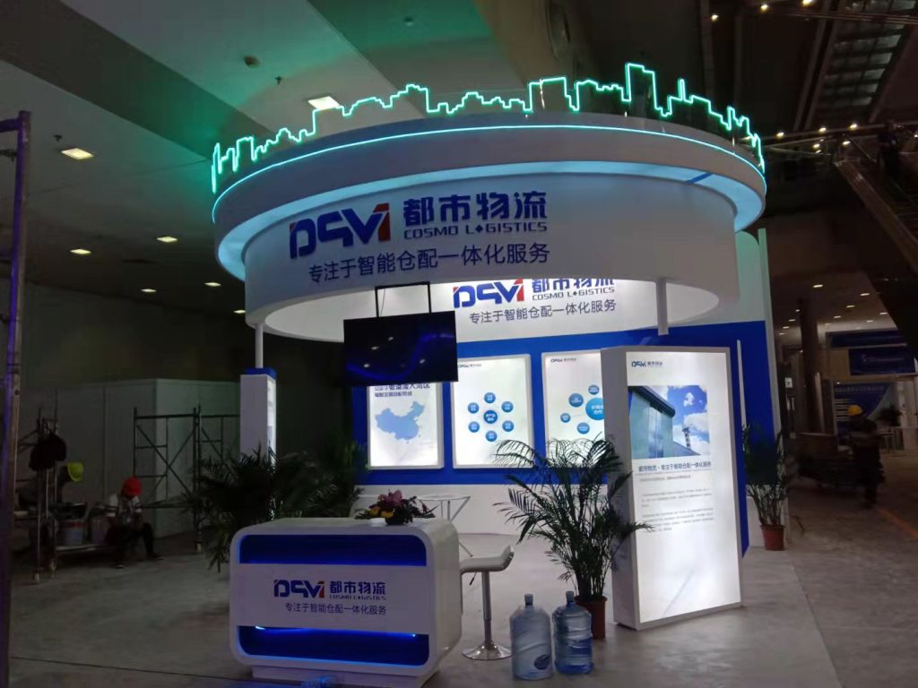 A white and blue trade show booth with a glowing green skyline graphic, featuring a large screen, multiple display panels, and potted plants.