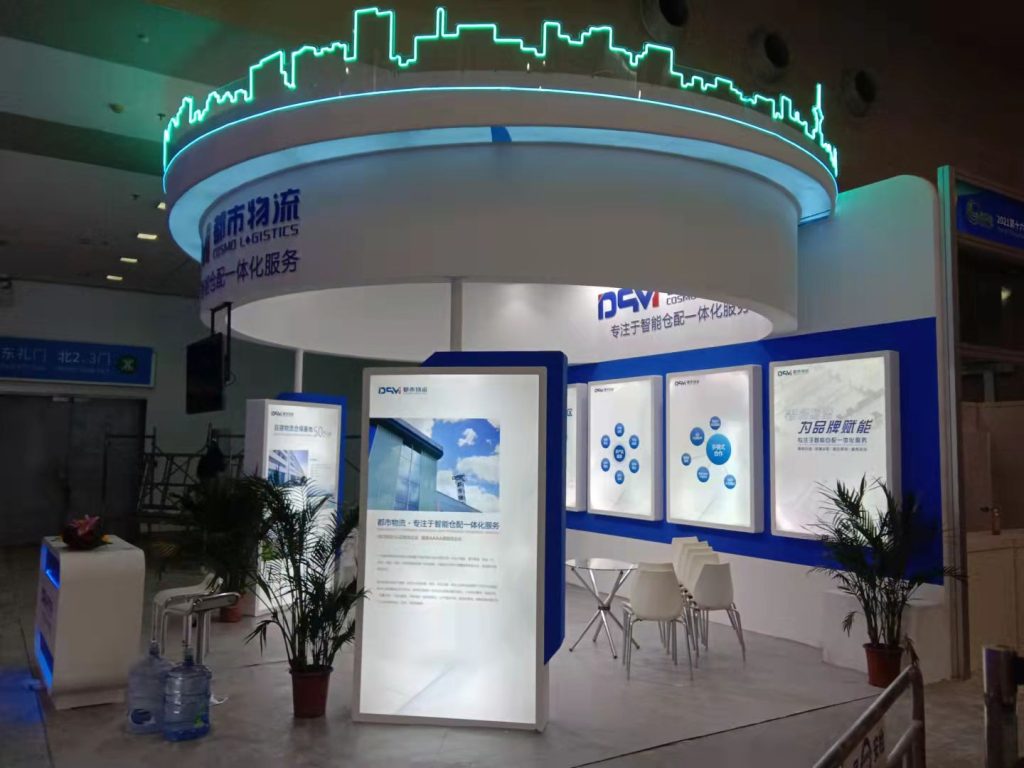 An angled view of a white and blue trade show booth with several illuminated display panels, a table with chairs, and potted plants.