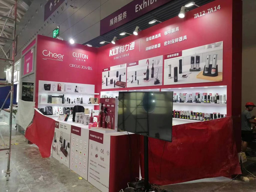 Red and white exhibition booth displaying kitchen gadgets on shelves, with a monitor in front. Red construction fabric partially covers the lower area.