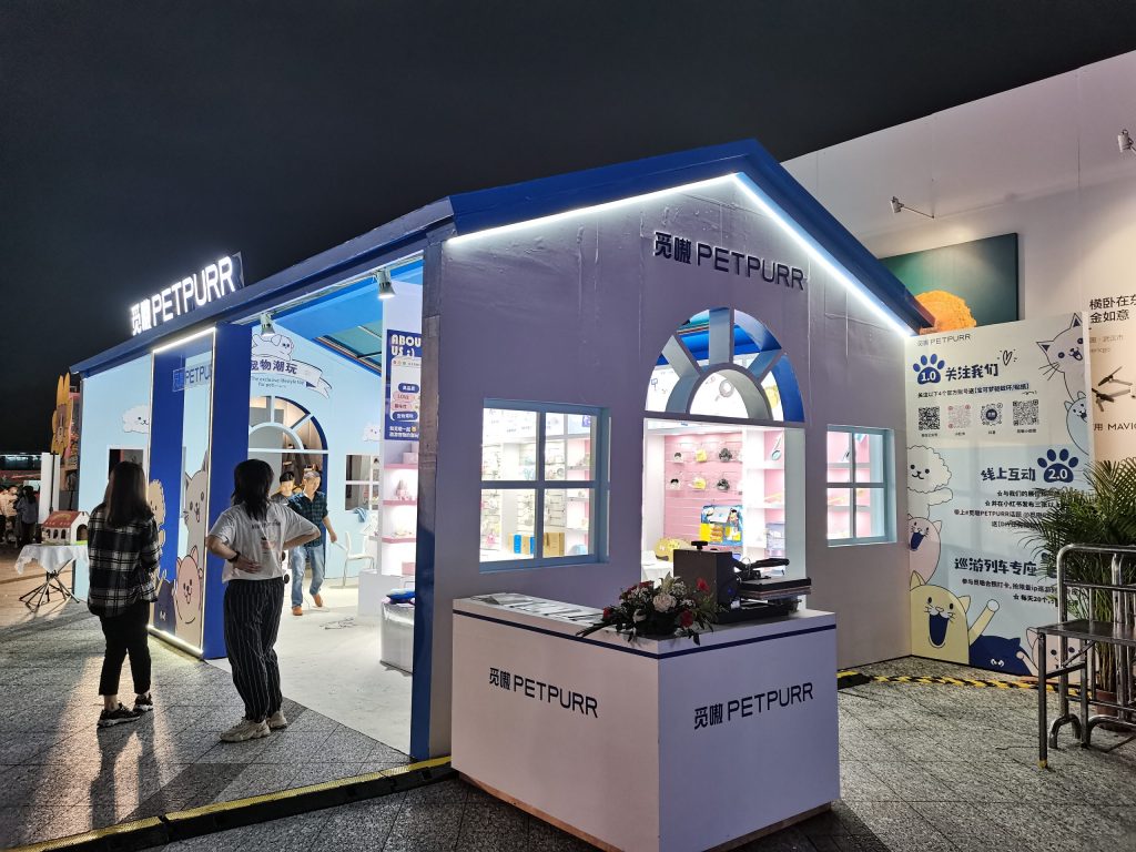 Two people stand outside a brightly lit house-shaped booth at an event. The booth features windows, a counter, and cartoon animal decorations.
